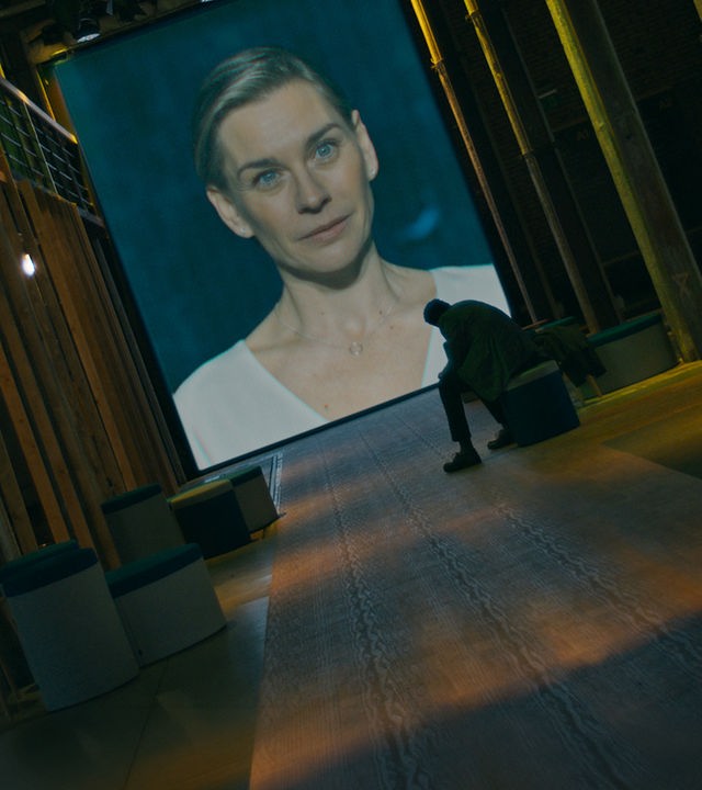 "Concordia - Tödliche Utopie (6) - Kein Weg zurück": Juliane Ericksen (Christiane Paul) ist auf eine Leinwand projiziert. Davor sitzt Noah Ericksen (Steven Sowah) in einem dunklen Raum und sieht zu.