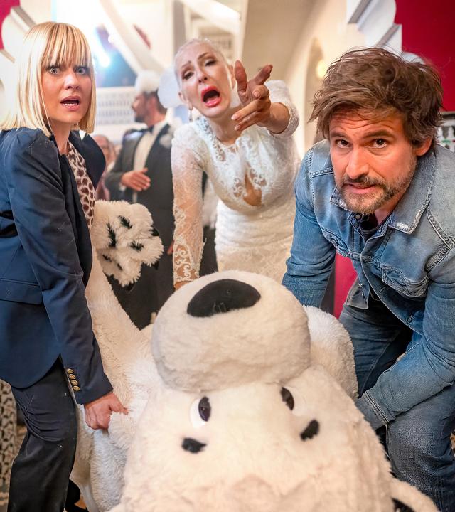 Julianes (Désirée Nick, M.) Hochzeitsfeier läuft vollkommen aus dem Ruder, als Nina (Henrike Fehrs, l.) und Tom (Tom Beck) versuchen, die Leiche im Eisbärkostüm aus der Location zu schaffen.