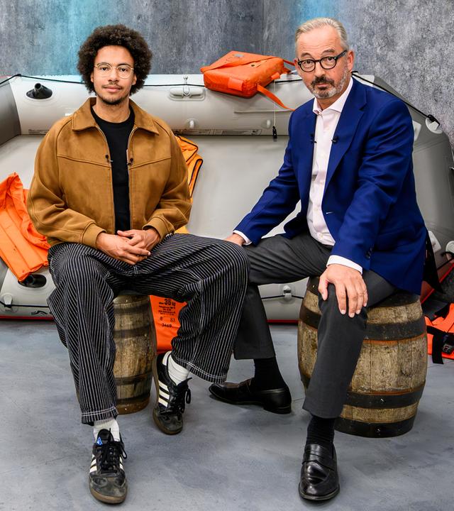  Luis Bobga und Jan Fleischhauer sitzen im Studio auf Holzfässern vor einem grauen Schlauchboot. 