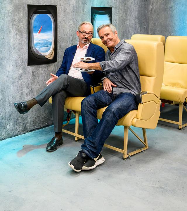 Jan Fleischhauer und Hannes Jaenicke sitzen nebeneinander auf gelben Flugzeugsitzen vor einer grauen Studiowand. Fleischhauer trägt ein blaues Sakko und eine graue Hose, Jaenicke ein graues Hemd, Jeans und schwarze Sportschuhe. An der Wand sind zwei nachgebildete Flugzeugfenster mit Wolken und einem Flugzeugflügel zu sehen. Rechts steht ein Servierwagen mit einer Flasche im Kühler und mehreren Gläsern.