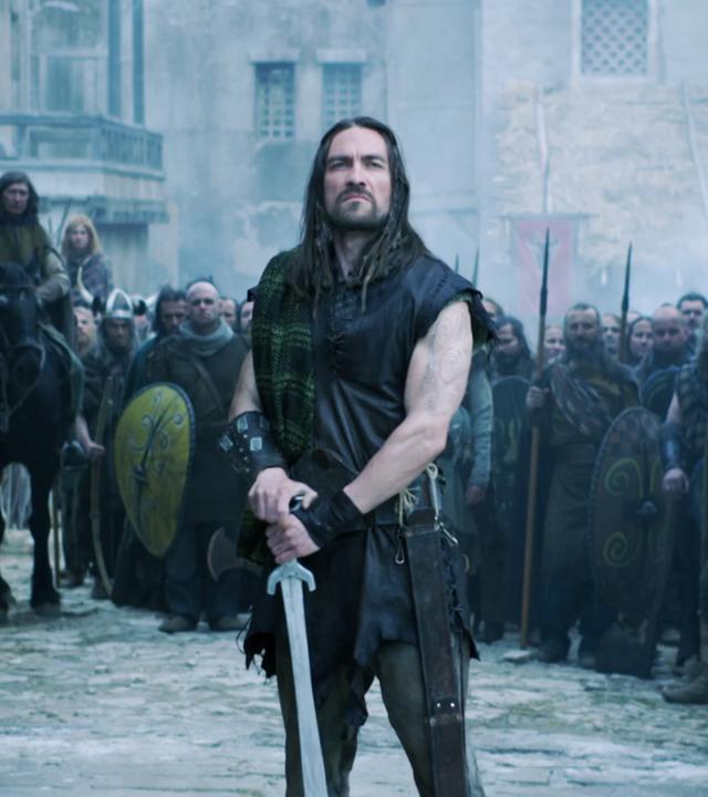 Ein Kelte mit Schwert steht in den Toren einer Stadt. Hinter ihm haben sich weitere Kämpfer versammelt.