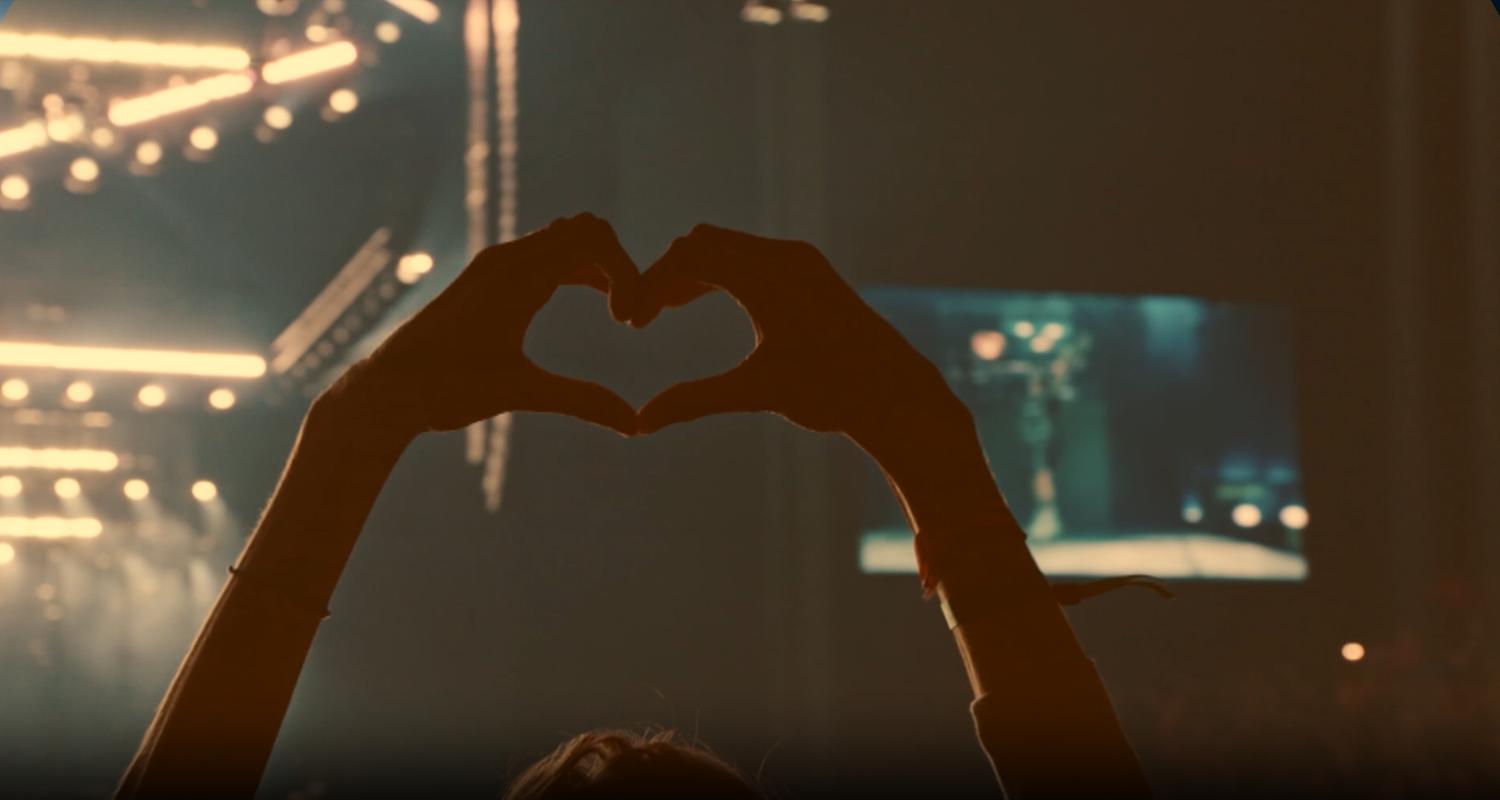 Hochgestreckte Hände bilden ein Herz vor einem verschwommenen Konzerthintergrund.