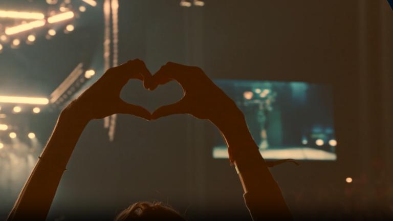 Hochgestreckte Hände bilden ein Herz vor einem verschwommenen Konzerthintergrund.