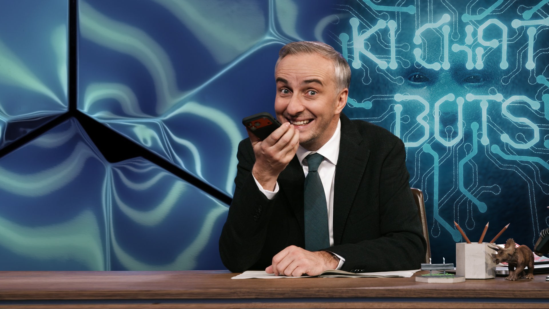 Jan Böhmermann sitzt am Schreibtisch und spricht in sein Smartphone. Rechts von ihm im Hintergrund steht "KI CHAT BOTS", hinter dem Schriftzug ist schemenhaft ein künstlich gestaltetes Gesicht abgebildet.