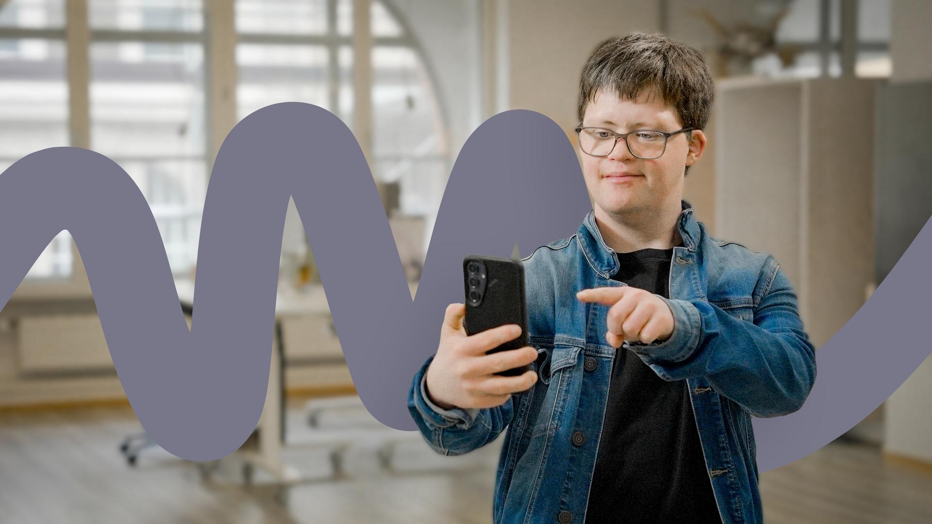 Arthur Hackenthal steht in einem hellen Büroraum und hält sein Smartphone in der rechten Hand. Er zeigt mit dem Zeigefinger auf das Gerät und lächelt in die Kamera. Er trägt eine Jeansjacke über einem schwarzen T-Shirt. Im Hintergrund ist ein großes Rundbogenfenster zu sehen.