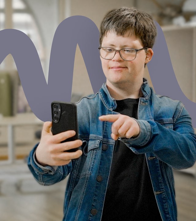 Arthur Hackenthal steht in einem hellen Büroraum und hält sein Smartphone in der rechten Hand. Er zeigt mit dem Zeigefinger auf das Gerät und lächelt in die Kamera. Er trägt eine Jeansjacke über einem schwarzen T-Shirt. Im Hintergrund ist ein großes Rundbogenfenster zu sehen.