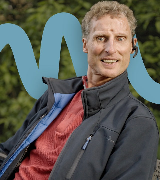 Andreas Loose sitzt mittig im Bild auf einer Bank. Er trägt eine dunkelblaue Jacke und einen roten Pullover. In den Händen hält er seinen Blindenstock. Er trägt sein Headset. Man sieht nur seinen Oberkörper. Er grinst in die Kamera. 