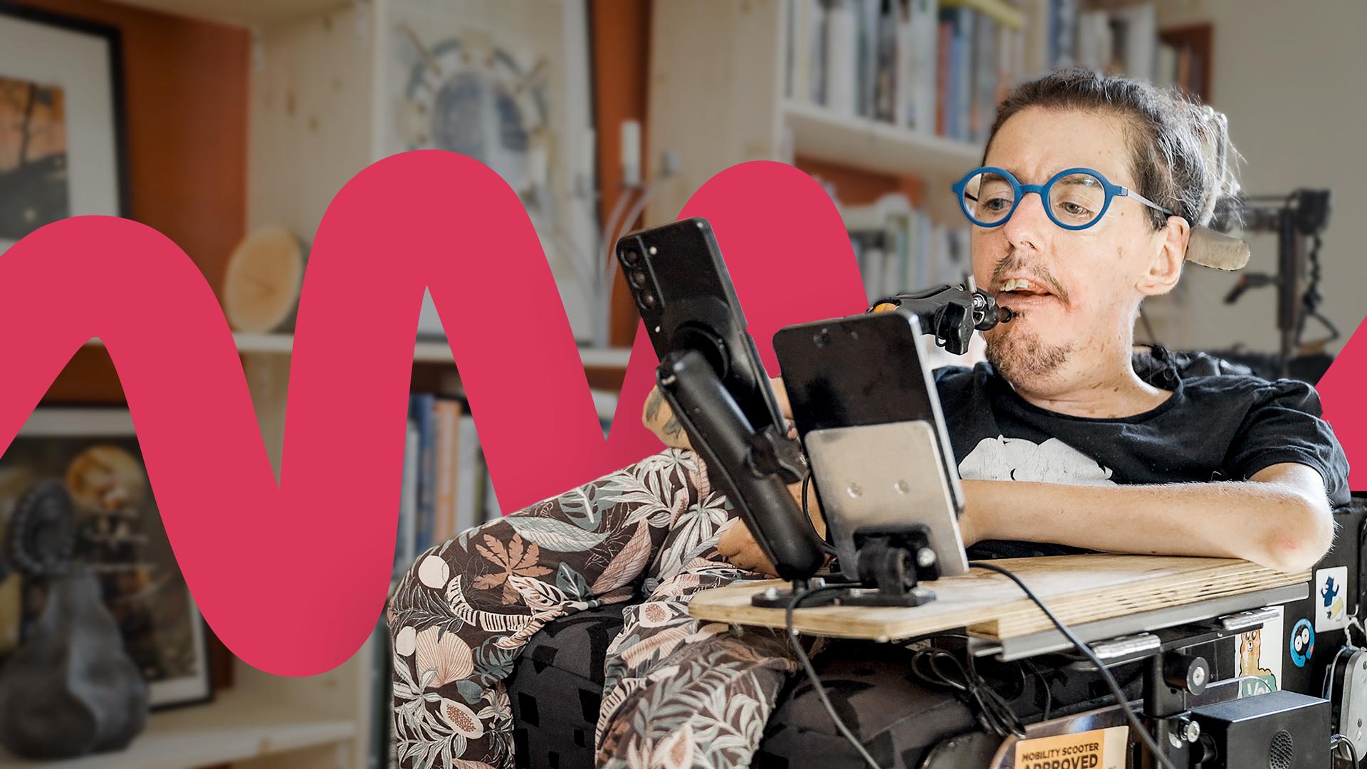 Christian Bayerlein sitzt in seinem Rollstuhl und schaut vor sich auf ein Smartphone. Er trägt ein schwarzes T-Shirt und eine blaue, runde Brille. Im Hintergrund ist ein Bücherregal voll mit Büchern und Kunst zu sehen.