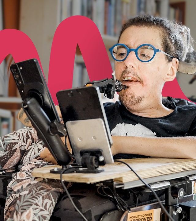 Christian Bayerlein sitzt in seinem Rollstuhl und schaut vor sich auf ein Smartphone. Er trägt ein schwarzes T-Shirt und eine blaue, runde Brille. Im Hintergrund ist ein Bücherregal voll mit Büchern und Kunst zu sehen.