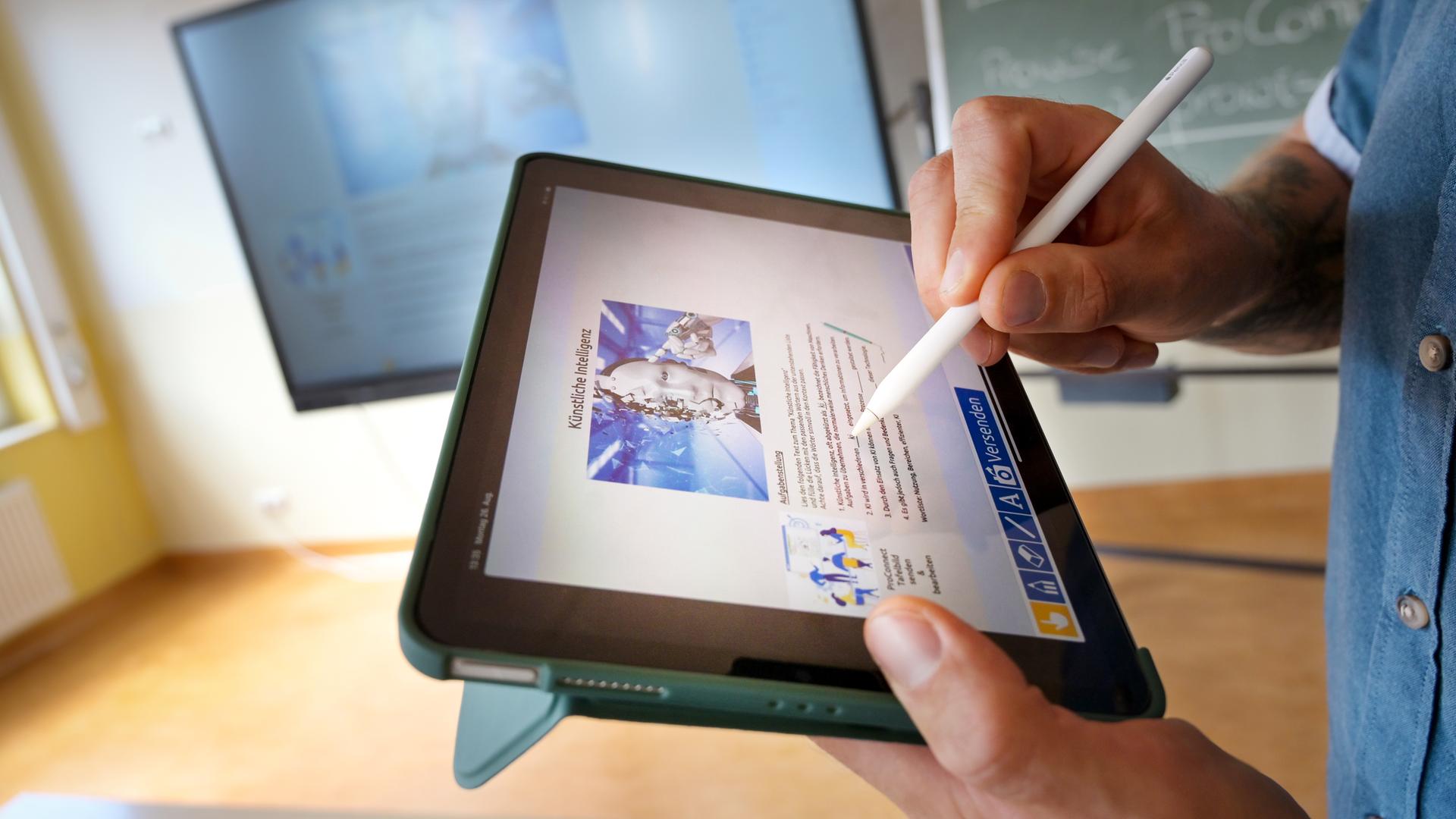 Lehrperson hält Tablet in der Hand, im Hintergrund sind zwei Schultafeln zu sehen.