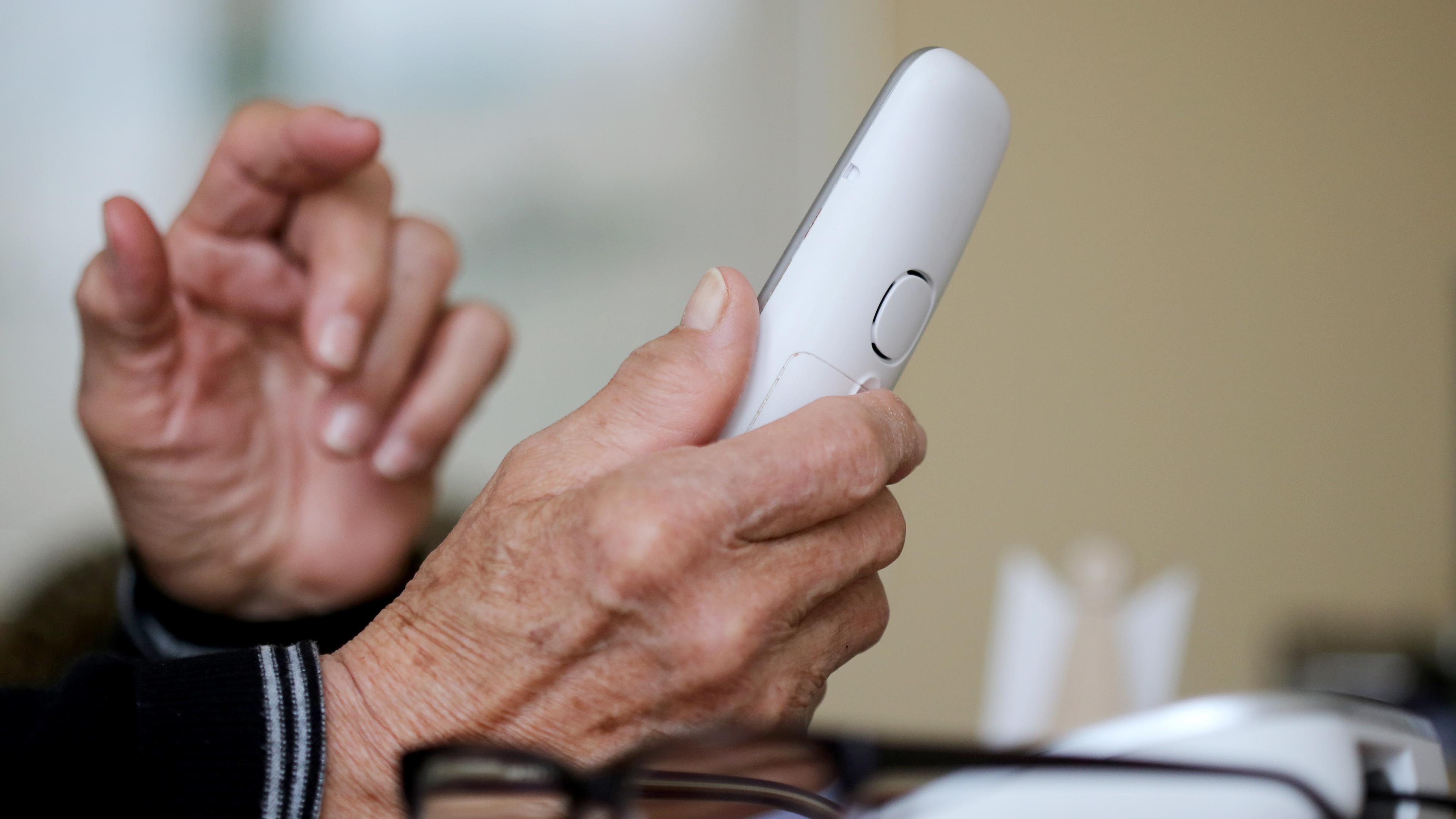 Die Hände einer älteren Person sind zu sehen, in der rechten Hand wird ein weißer Telefonhörer gehalten.