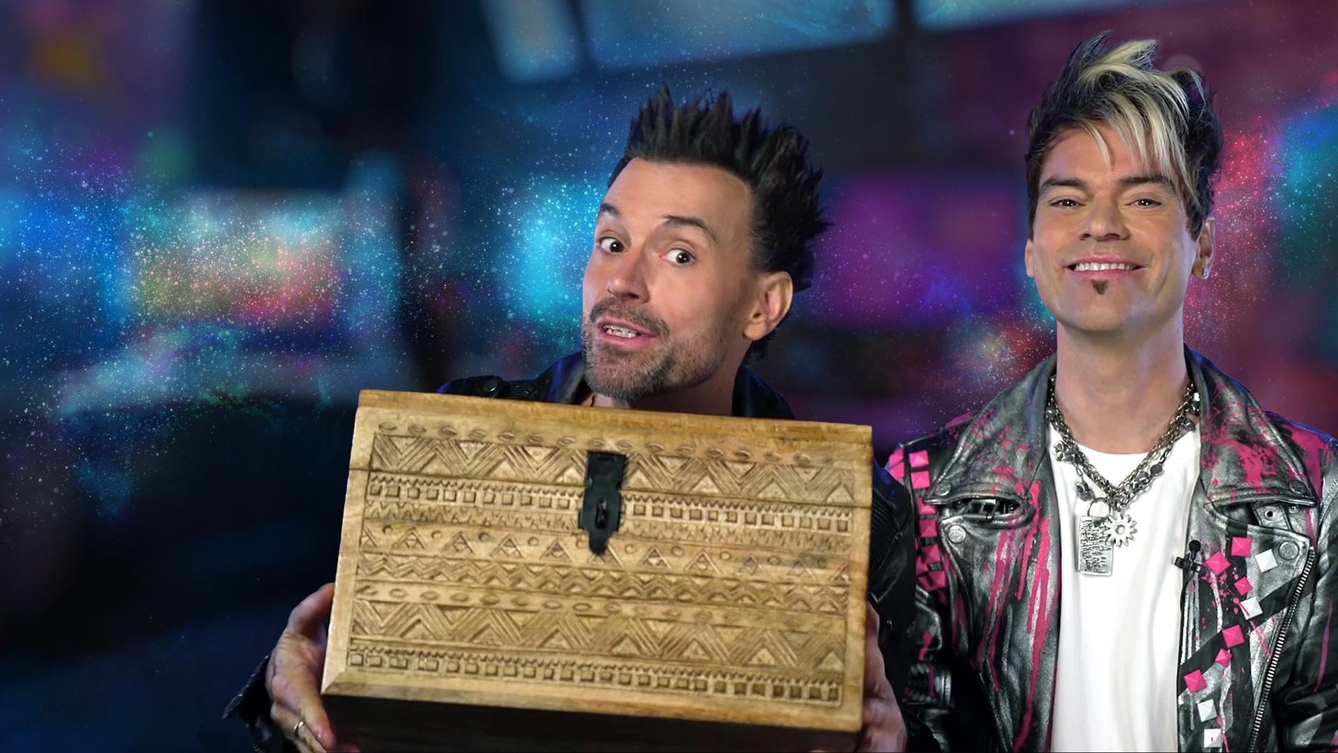 Die Ehrlich Brothers sind vor einem bunten Hintergrund zu sehen, Chris lächelt in die Kamera und Andreas hat eine Schatzkiste in der Hand.