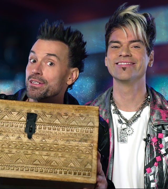 Die Ehrlich Brothers sind vor einem bunten Hintergrund zu sehen, Chris lächelt in die Kamera und Andreas hat eine Schatzkiste in der Hand.