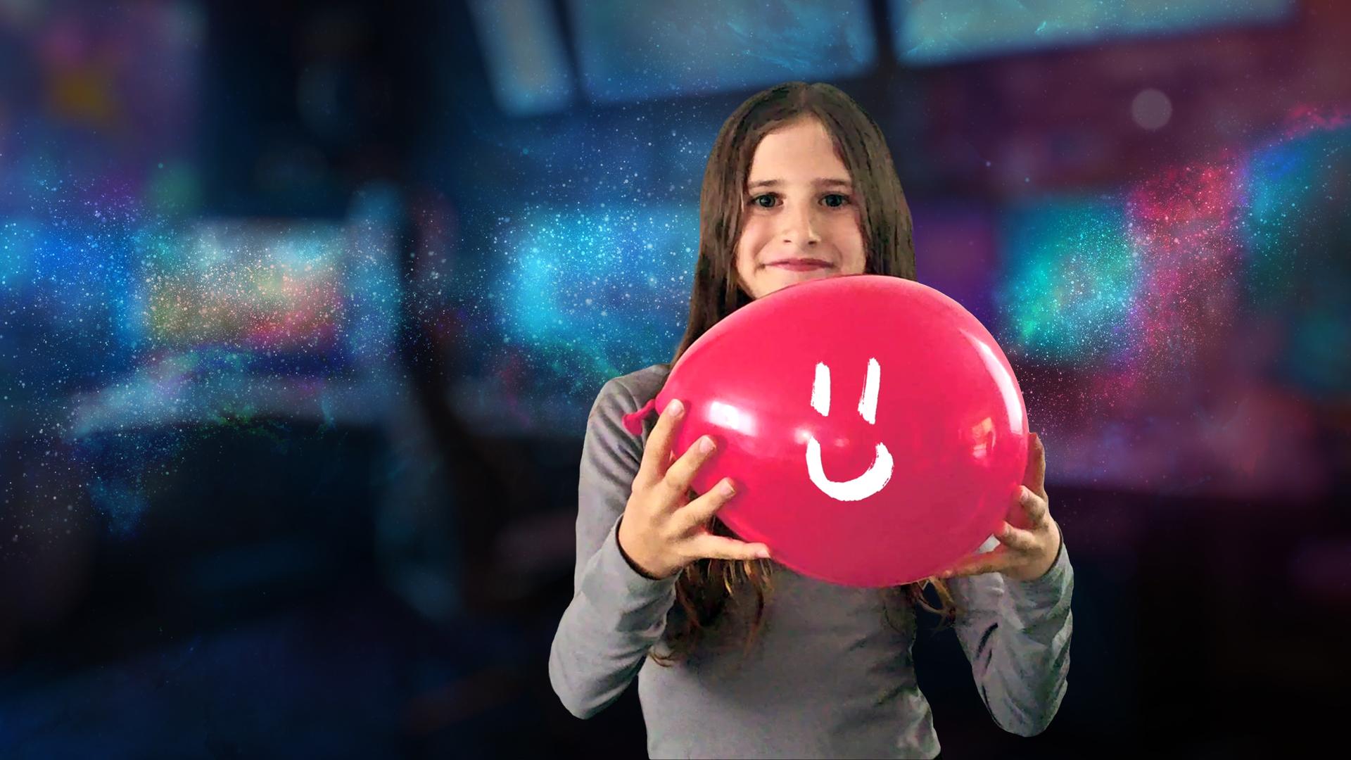 Ein kleines Mädchen schaut lächelnd in die Kamera und hält einen roten Ballon in der Hand, auf der ein weißes Lächeln gezeichnet ist.