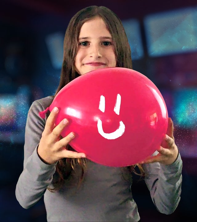 Ein kleines Mädchen schaut lächelnd in die Kamera und hält einen roten Ballon in der Hand, auf der ein weißes Lächeln gezeichnet ist.