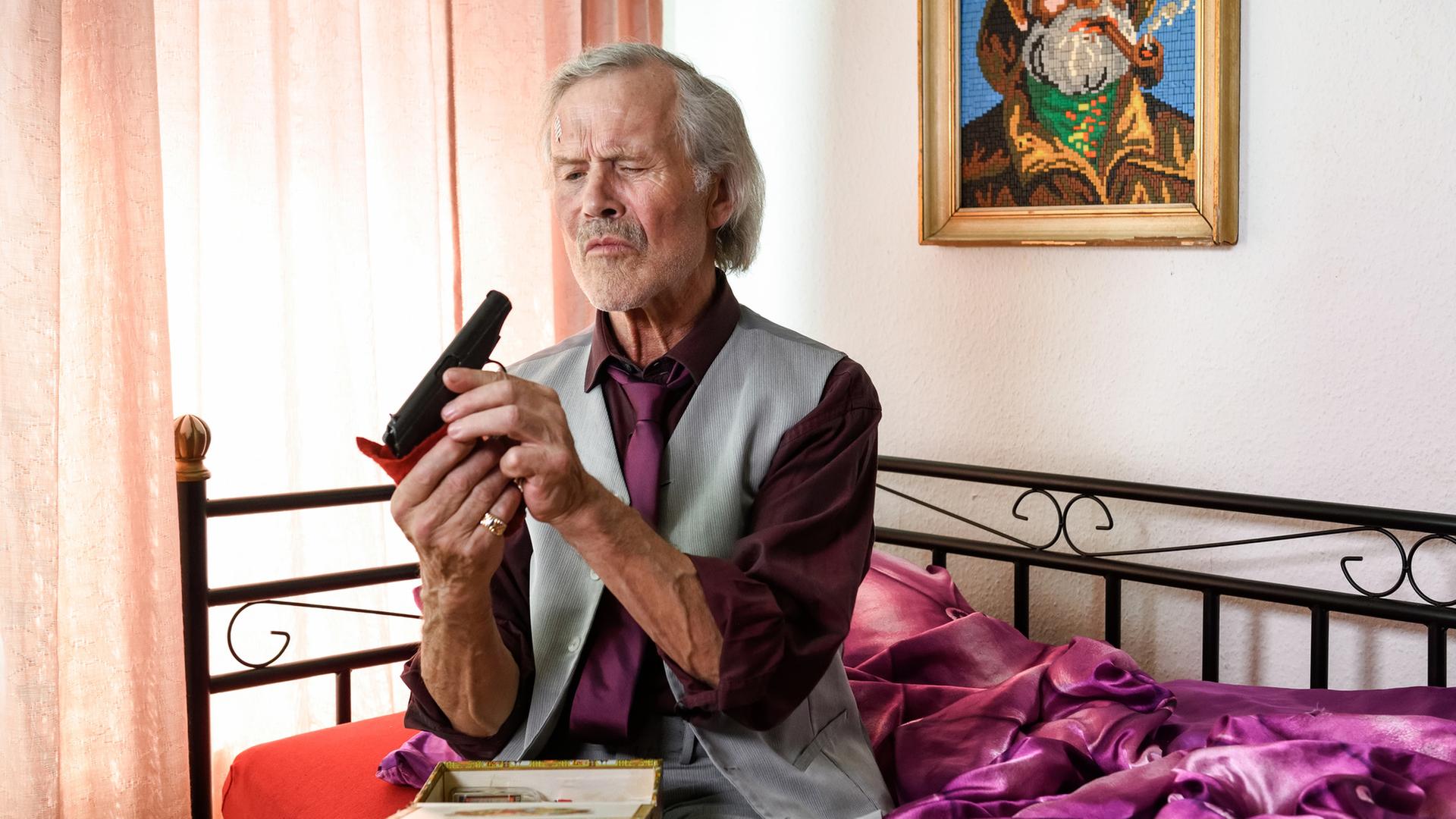 "Notruf Hafenkante - Kiezlösung": "Dandy" Hartmann (Heinrich Giskes) sitzt auf einem Bett. Er hält eine Pistole in den Händen, die er reinigt. Auf seinem Schoß steht ein geöffnetes Kästchen.