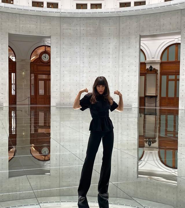  In einem runden Raum mit hohen Betonwänden und einem spiegelnden Boden steht eine Frau mit langen, dunklen Haaren und hebt beide Arme an, als würde sie ihre Muskeln präsentieren. Sie trägt ein komplett schwarzes Outfit, bestehend aus einer taillierten Bluse mit kurzen Ärmeln und einer weiten Hose. Ihr Blick ist selbstbewusst und direkt in die Kamera gerichtet. Der Boden reflektiert die Umgebung, wodurch es scheint, als würde der Raum nach unten hin weitergehen. Durch zwei große, rechteckige Öffnungen in den Betonwänden sind elegante, historische Elemente wie Holzvertäfelungen, Rundbögen und eine große Wanduhr sichtbar.