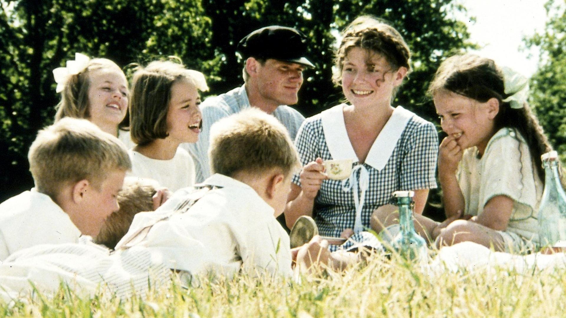 Die Kinder von Bullerbü sitzen gemeinsam mit der Magd Agda und dem Knecht Oskar bei sommerlichem Wetter auf einer grünen Wiese und lachen.