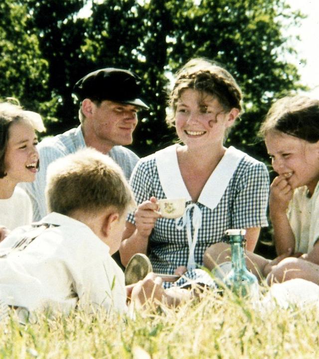 Die Kinder von Bullerbü sitzen gemeinsam mit der Magd Agda und dem Knecht Oskar bei sommerlichem Wetter auf einer grünen Wiese und lachen.