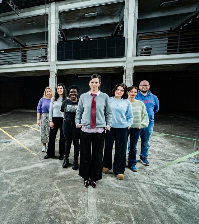 Es stehen sieben Menschen nebeneinander auf einem Boden, der ein schachbrettartiges Muster hat. Alle blicken in die Richtung des Betrachters.