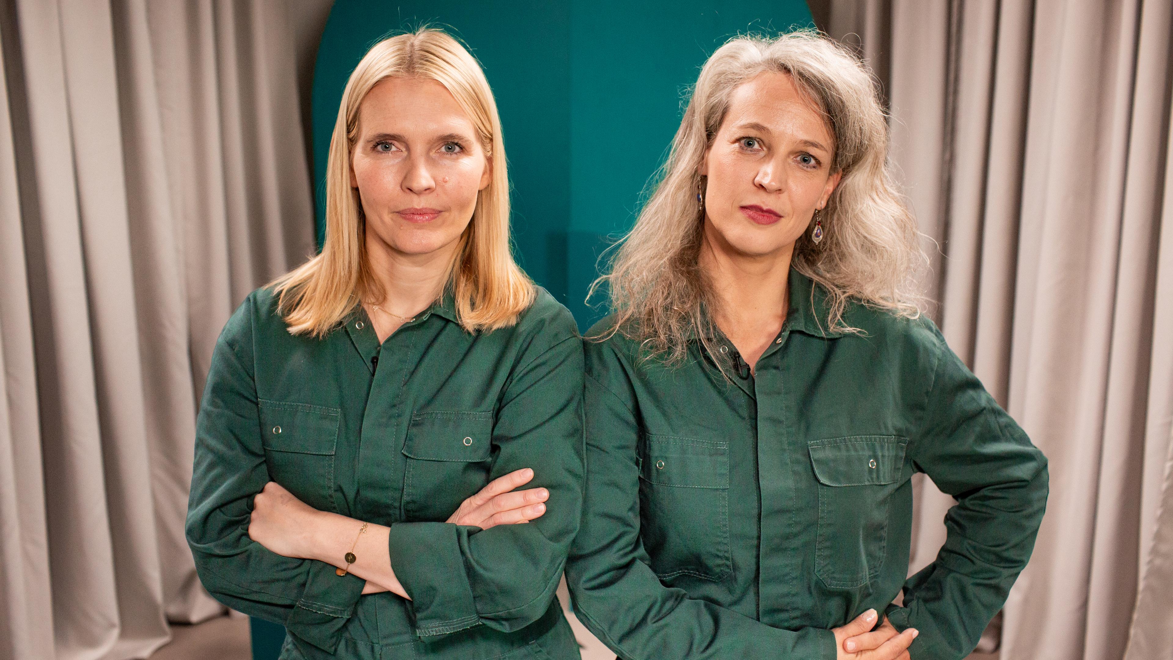 Zwei blonde, einen grünen Overall tragende Frauen stehen mit verschränkten Armen nebeneinander.