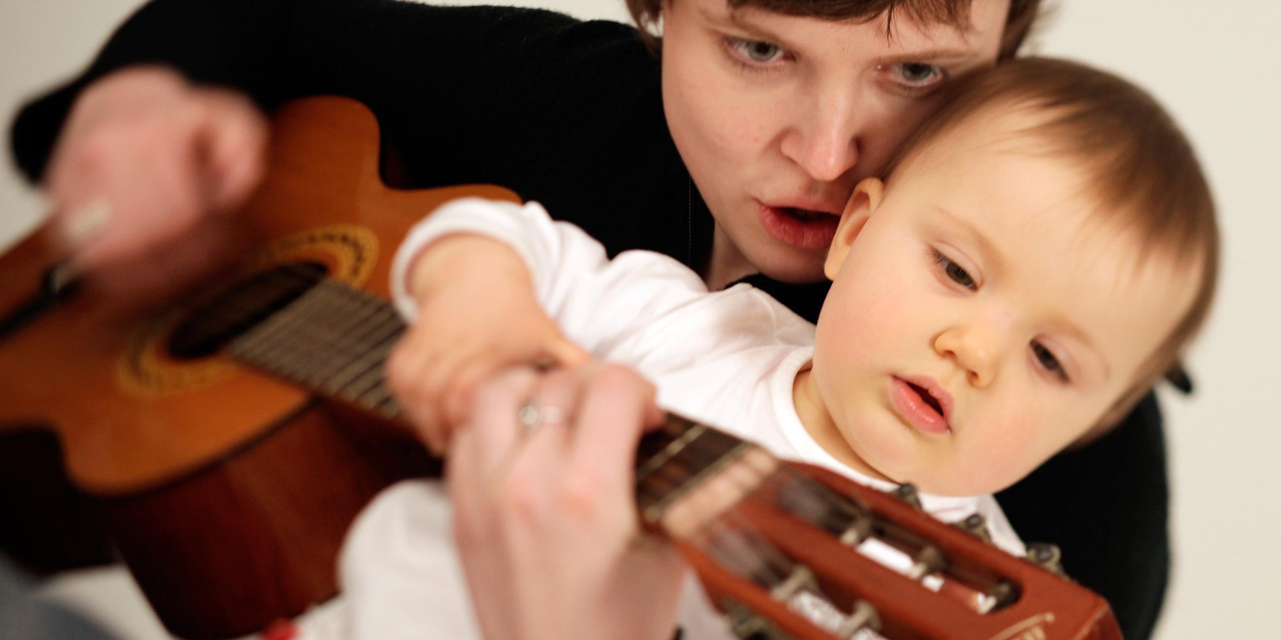 Eine Mutter hält ihr Kleinkind und eine Gitarre und spielt darauf.