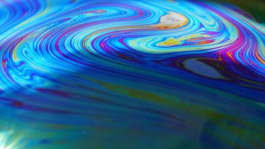 Abstraktes Bild: Schillernde Farbverläufe in Blau, Violett und Gelb fließen ineinander und erzeugen ein abstraktes, wellenartiges Muster. Die Darstellung erinnert an die Oberfläche einer Seifenblase in Nahaufnahme.