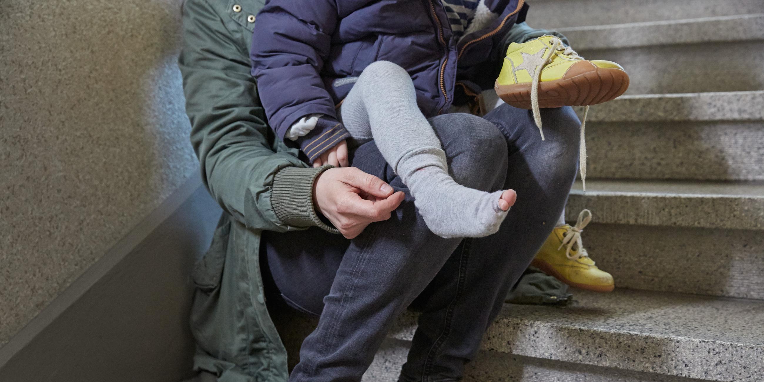 Eine Mutter sitzt im Treppenhaus und zieht ihrem Kind, in dessen Strumpf sich ein Loch befindet, die Schuhe an. (Symbolbild)