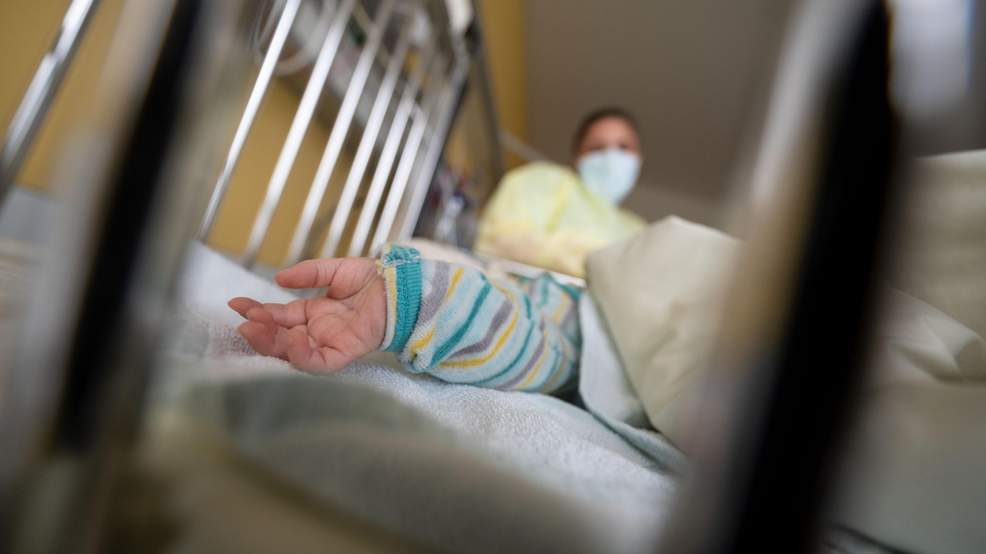 Die Hand eines Kindes in einem Krankenbett, im verschwommenen Hintergrund sieht man eine Krankenpflegerin/Ärztin