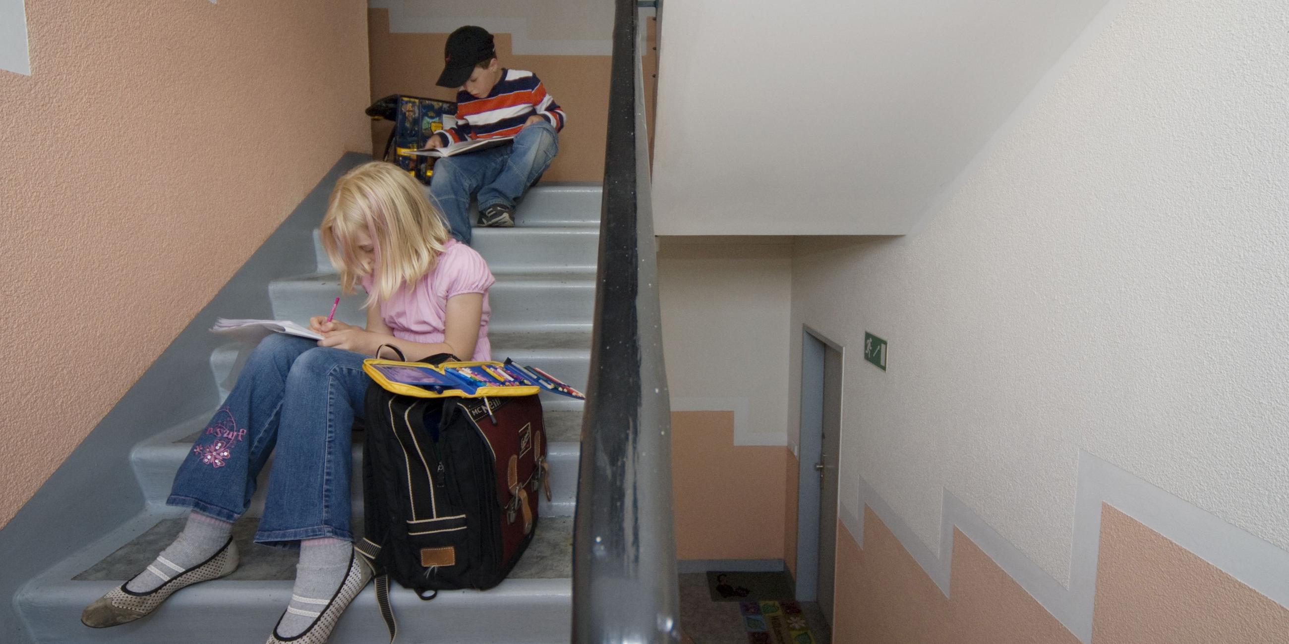 Zwei Kinder mit Schulranzen warten in einem Hausflur auf der Treppe.