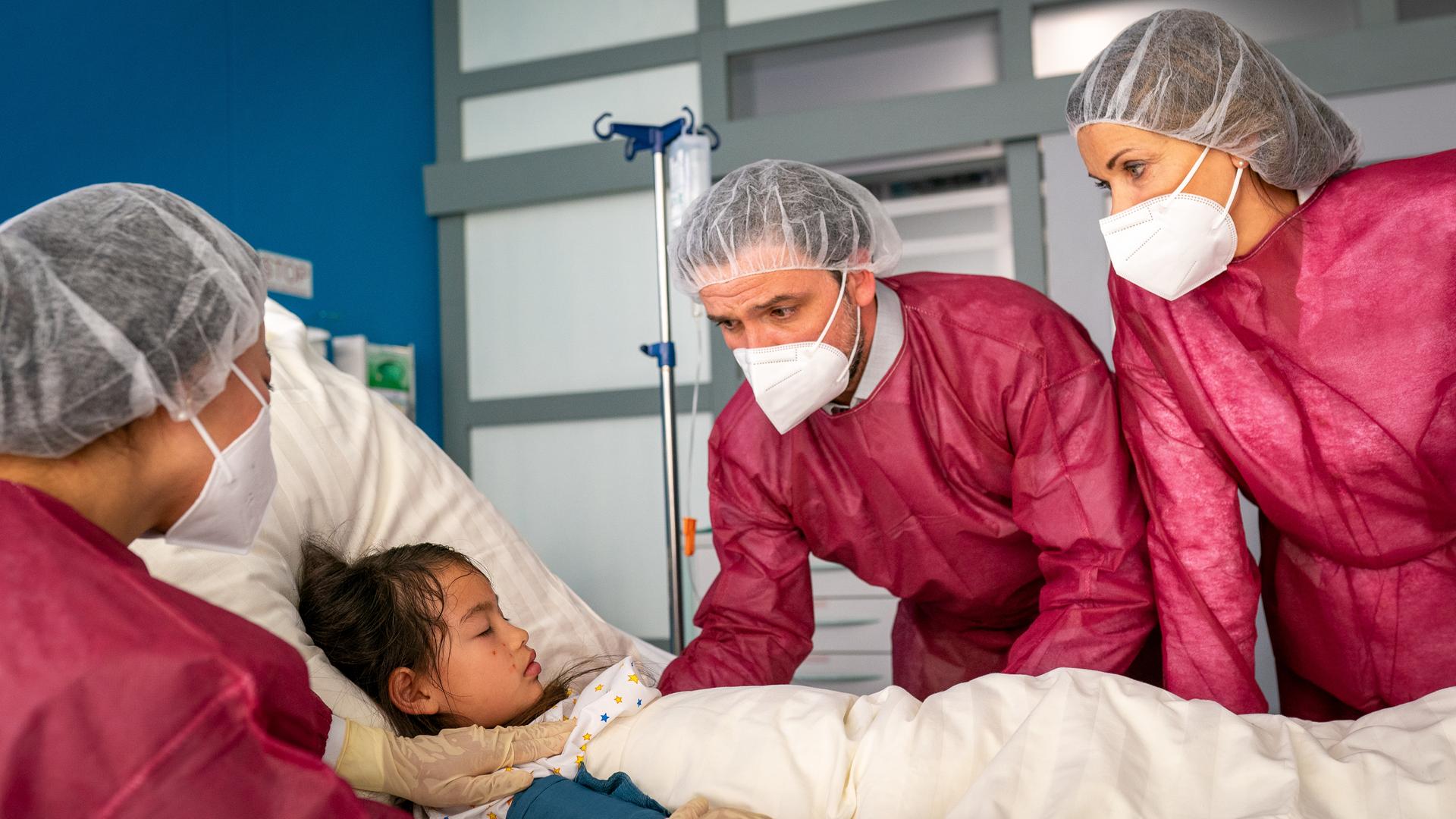 Nora (Kotti Yun), Roman (Sascha Goepel) und Dr. Jonas (Gerit Kling) stehen um das Krankenbett von Yella (Loretta Rühling). Alle tragen rote Schutzkittel, Masken, Handschuhe und Klipphauben. Alle drei sehen Yella besorgt an.