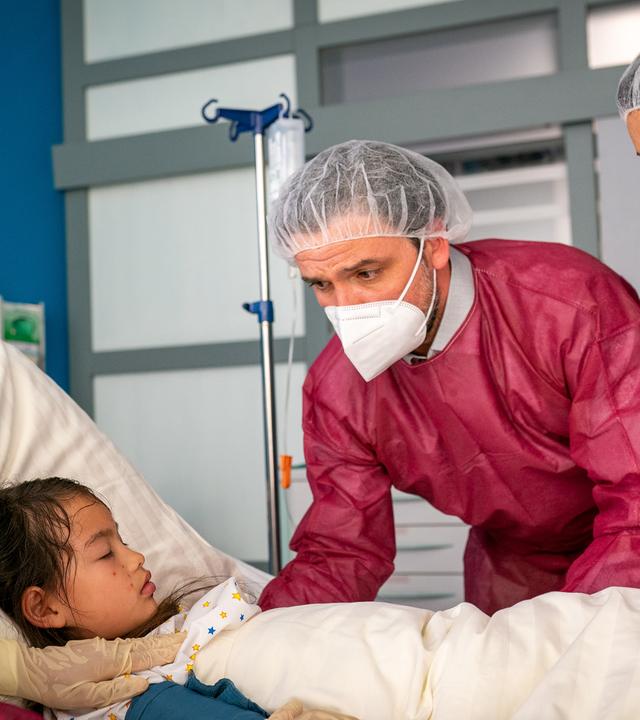 Nora (Kotti Yun), Roman (Sascha Goepel) und Dr. Jonas (Gerit Kling) stehen um das Krankenbett von Yella (Loretta Rühling). Alle tragen rote Schutzkittel, Masken, Handschuhe und Klipphauben. Alle drei sehen Yella besorgt an.