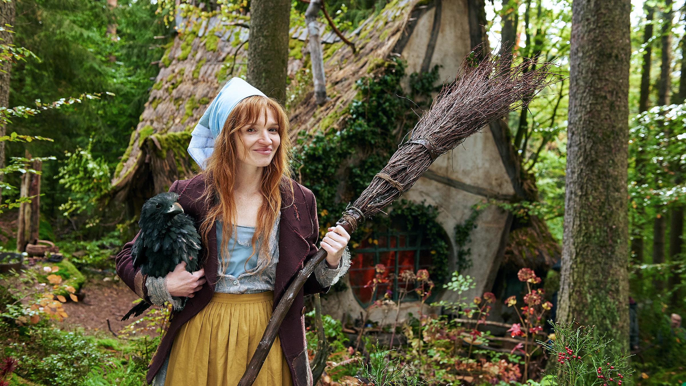 "Die kleine Hexe": Die kleine Hexe (Karoline Herfurth) steht lächelnd vor ihrem Hexenhaus im Wald. Im rechten Arm hält sie ihren Raben Abrayas, in der linken Hand ihren Hexenbesen.