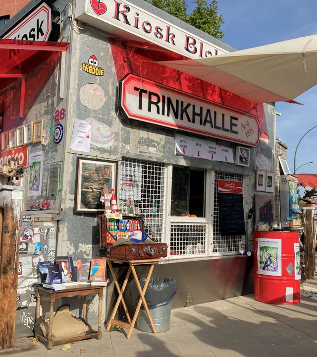 Das Bild zeigt einen Kiosk mit dem Namen „Trinkhalle“, der an einer Straßenecke steht. Über dem Schalter hängt ein Schild mit der Aufschrift „Trinkhalle Seit 1876“. Die Fassade des Kiosks ist abgenutzt und mit zahlreichen Stickern und Graffiti bedeckt. Vor dem Kiosk stehen einige Bücher auf einem kleinen Tisch, daneben verschiedene dekorative Elemente wie Bilder und andere Kunstgegenstände. Rechts im Bild sind einige Menschen unter einem Sonnenschirm zu sehen, die an Tischen stehen. 