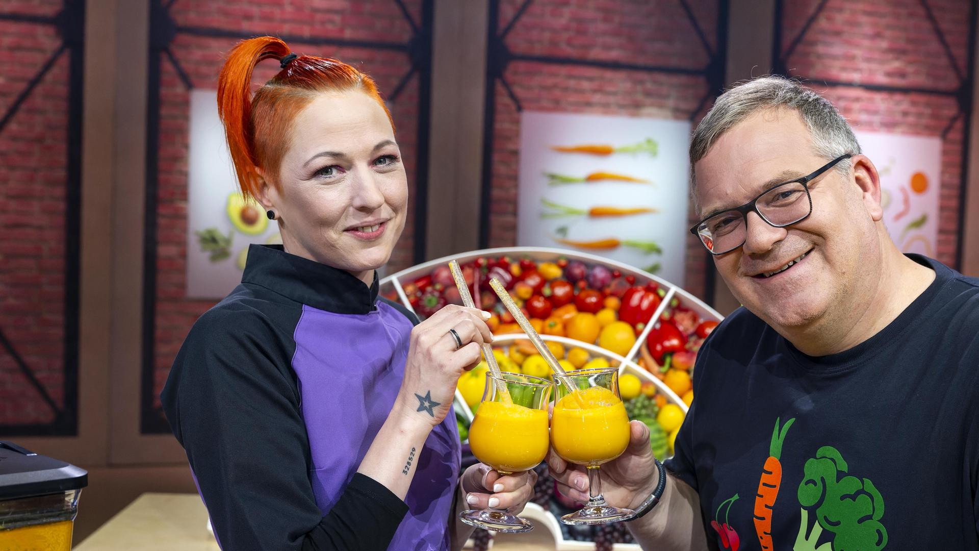 TV-Köchin Meta Hiltebrand (l.) hält in der linken Hand ein Glas Orangensaft. Mit der rechten Hand hält sie einen Strohhalm und prostet Elton zu, der ebefalls ein Glas Orangensaft hält.