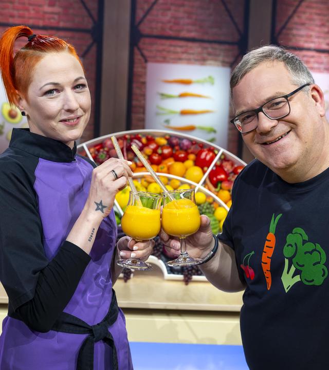 TV-Köchin Meta Hiltebrand (l.) hält in der linken Hand ein Glas Orangensaft. Mit der rechten Hand hält sie einen Strohhalm und prostet Elton zu, der ebefalls ein Glas Orangensaft hält.