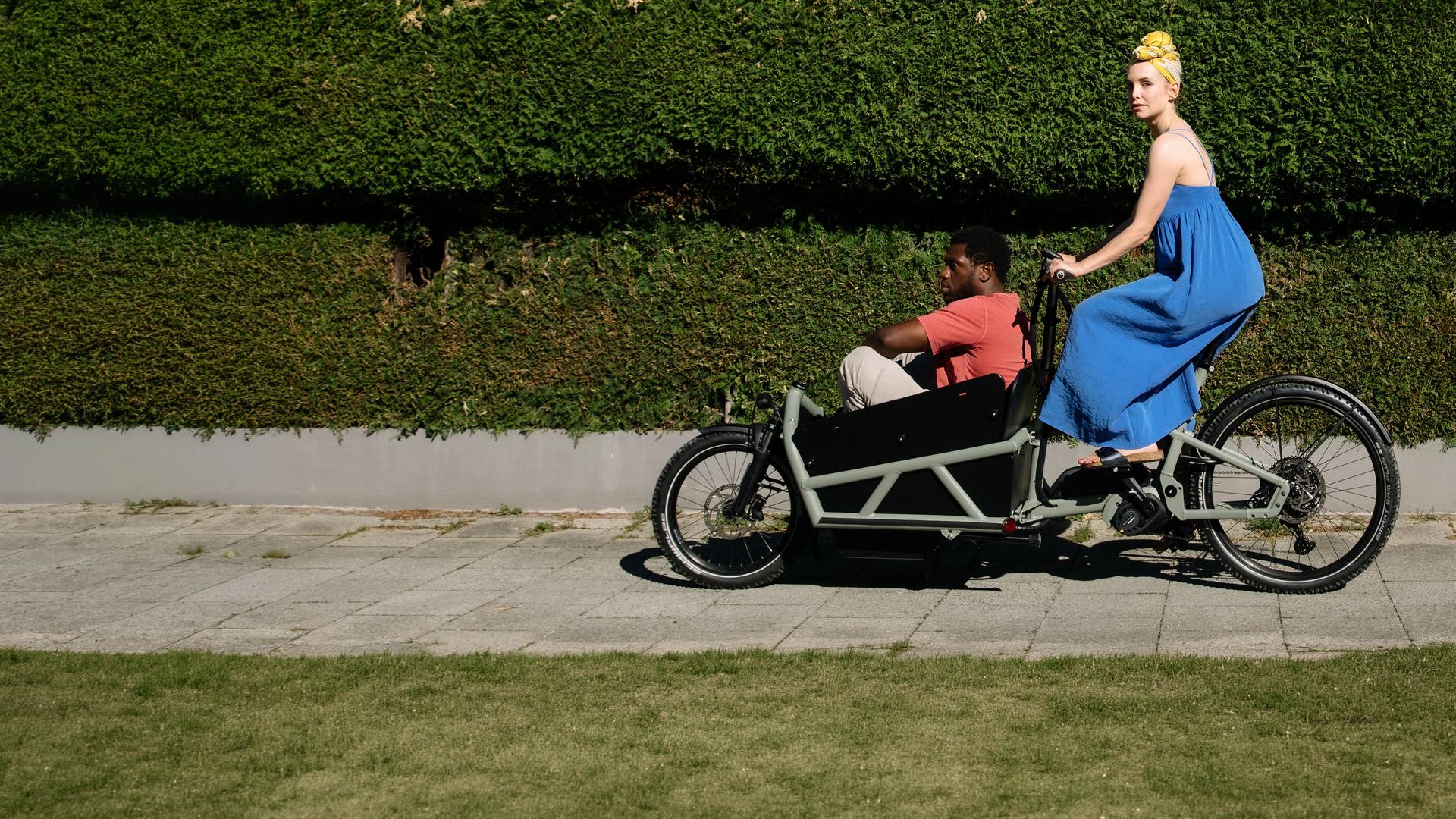 "I don't work here - Kitzbühel": Laura (Sina Martens) fährt in einem blauen Kleid auf einem schwarzen Lastenrad an einer grünen Hecke vorbei, hinter der ein weißes Haus mit zwei Fenstern zu sehen ist. Dawit (Akeem von Flodrop) sitzt mit angezogenen Beinen vorne im Lastenrad und lässt sich von Laura fahren, die zur Seite schaut.