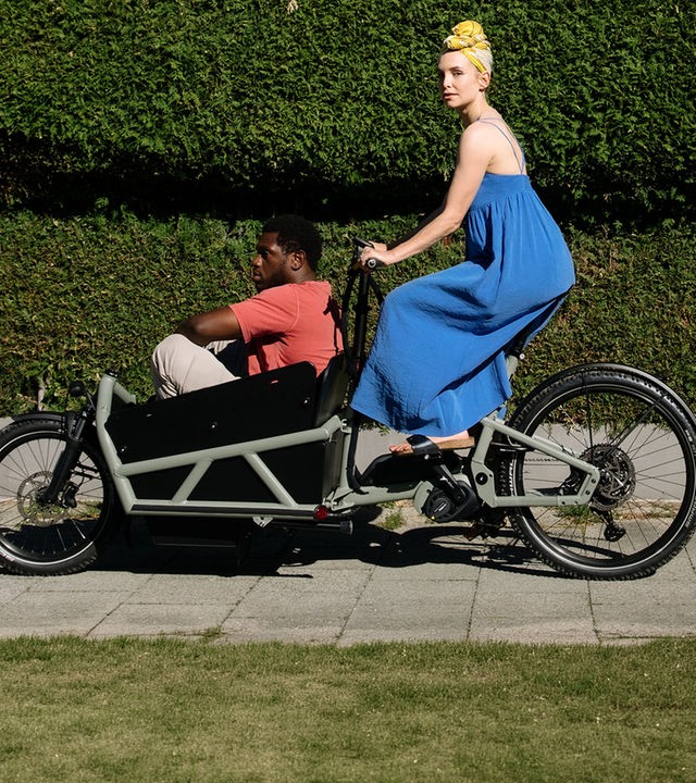 "I don't work here - Kitzbühel": Laura (Sina Martens) fährt in einem blauen Kleid auf einem schwarzen Lastenrad an einer grünen Hecke vorbei, hinter der ein weißes Haus mit zwei Fenstern zu sehen ist. Dawit (Akeem von Flodrop) sitzt mit angezogenen Beinen vorne im Lastenrad und lässt sich von Laura fahren, die zur Seite schaut.
