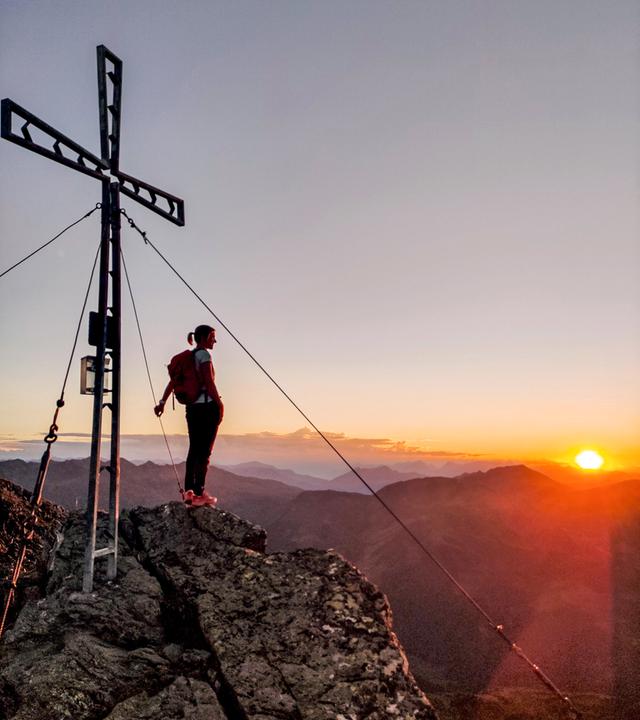 Frau steht in Wanderkleidung am Gipfelkreuz und betrachtet die Sonne über dem Panorama