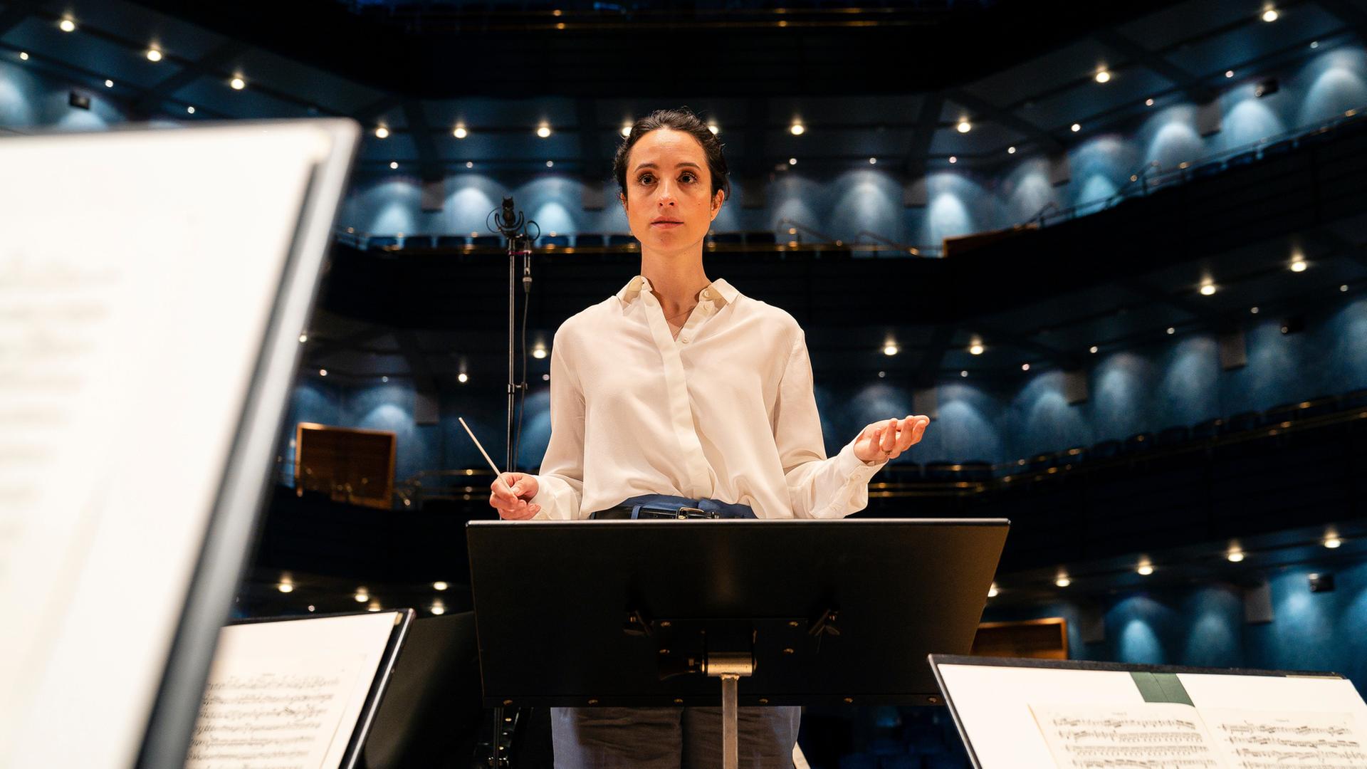 "Inga Lindström: Klang der Sehnsucht": Stine in weißer Bluse steht hinter einem Dirigentenpult, vor ihr stehen Notenständern. Sie hat beide Hände leicht angehoben, gleich gibt sie den Orchestermusikern den Einsatz.