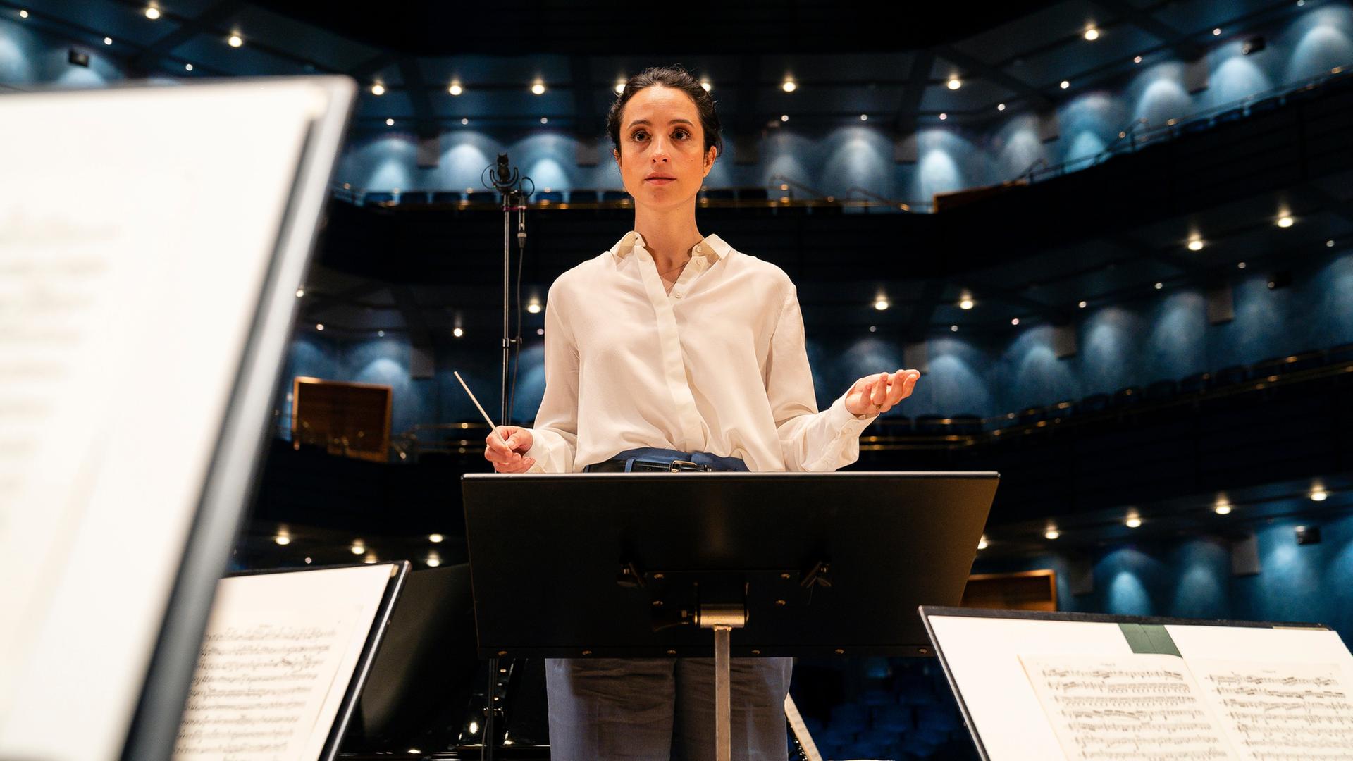"Inga Lindström: Klang der Sehnsucht": Stine in weißer Bluse steht hinter einem Dirigentenpult, vor ihr stehen Notenständern. Sie hat beide Hände leicht angehoben, gleich gibt sie den Orchestermusikern den Einsatz.
