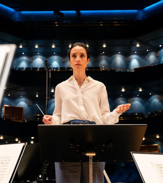 "Inga Lindström: Klang der Sehnsucht": Stine in weißer Bluse steht hinter einem Dirigentenpult, vor ihr stehen Notenständern. Sie hat beide Hände leicht angehoben, gleich gibt sie den Orchestermusikern den Einsatz.