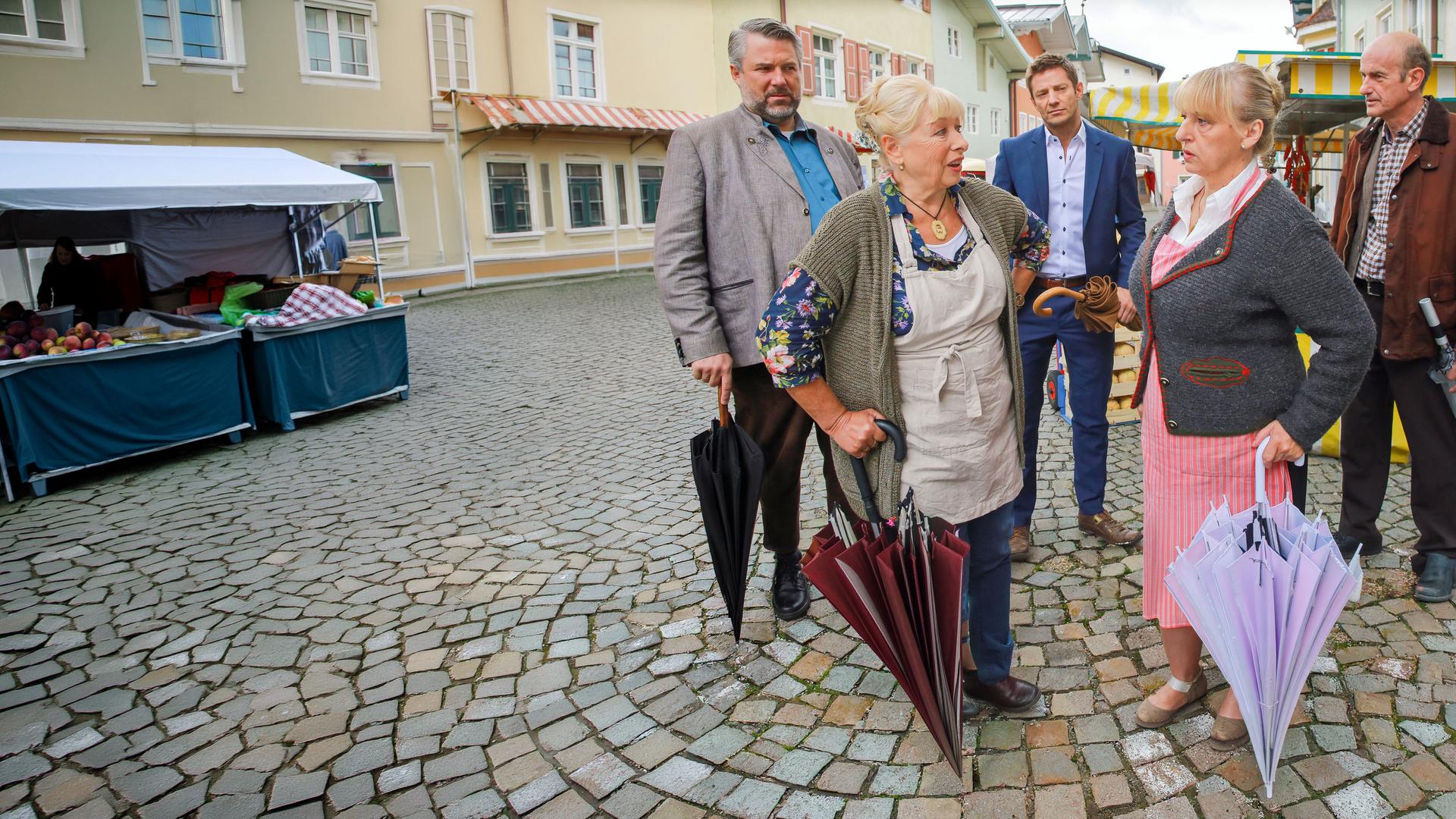 "Die Rosenheim-Cops - Klappe zu, Marktfrau tot": Die Kommissare Stadler (Dieter Fischer) und Hansen (Igor Jeftic) stehen auf der Straße vor Marktständen und betrachten die Standbetreiberinnen Marianne Widl (Erika Fischer-Laughlin) und Gertie Pointner (Johanna Bittenbinder), die sich unterhalten. Neben den Kommissaren steht Werner Kraisig (Johannes Herrschmann), der zu Stadler schaut.