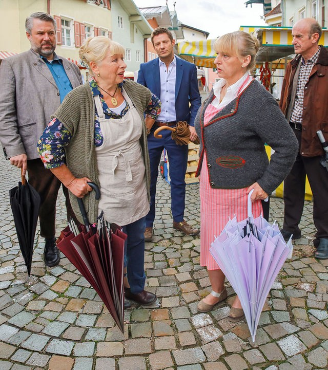 "Die Rosenheim-Cops - Klappe zu, Marktfrau tot": Die Kommissare Stadler (Dieter Fischer) und Hansen (Igor Jeftic) stehen auf der Straße vor Marktständen und betrachten die Standbetreiberinnen Marianne Widl (Erika Fischer-Laughlin) und Gertie Pointner (Johanna Bittenbinder), die sich unterhalten. Neben den Kommissaren steht Werner Kraisig (Johannes Herrschmann), der zu Stadler schaut.