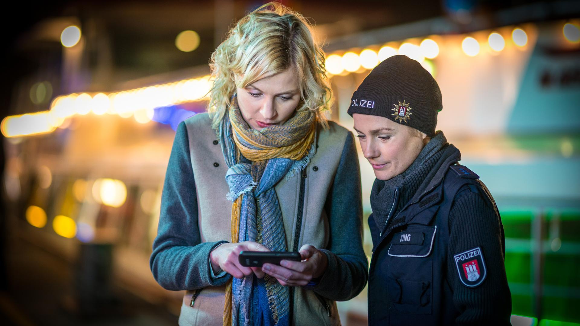"Notruf Hafenkante - Klassentreffen": Die Polizeibeamtin Franzi Jung (Rhea Harder-Vennewald) steht neben Melanie Hansen (Sanna Englund) und schaut auf deren Handy, in dem Melanie ihr etwas zeigt.