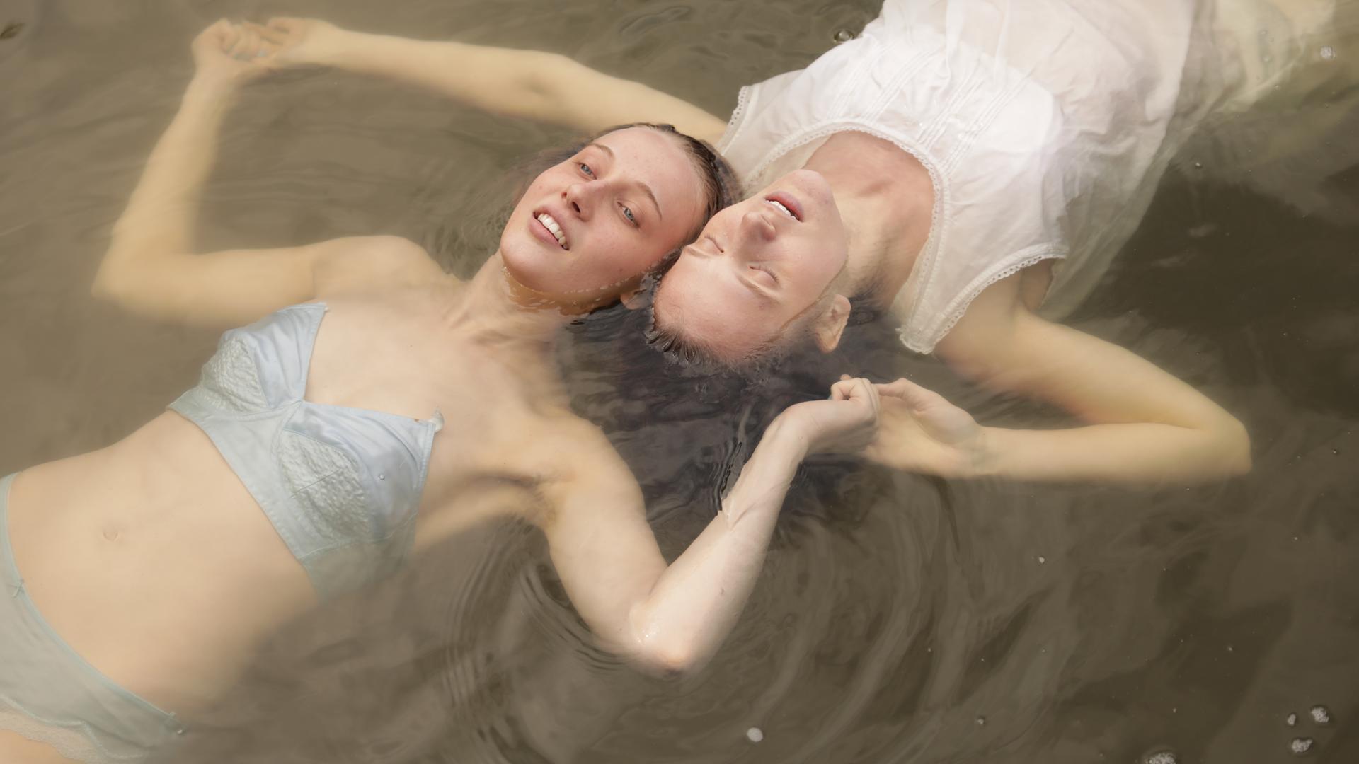 Zwei Mädchen schwimmen auf dem Rücken Kopf an Kopf in einem See und halten sich neben den Köpfen an den Händen. Das Mädchen vorne trägt blaue Uterwäsche und das andere Mädchen ein weißes Nachthemd.