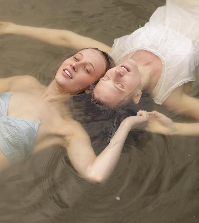 Zwei Mädchen schwimmen auf dem Rücken Kopf an Kopf in einem See und halten sich neben den Köpfen an den Händen. Das Mädchen vorne trägt blaue Uterwäsche und das andere Mädchen ein weißes Nachthemd.