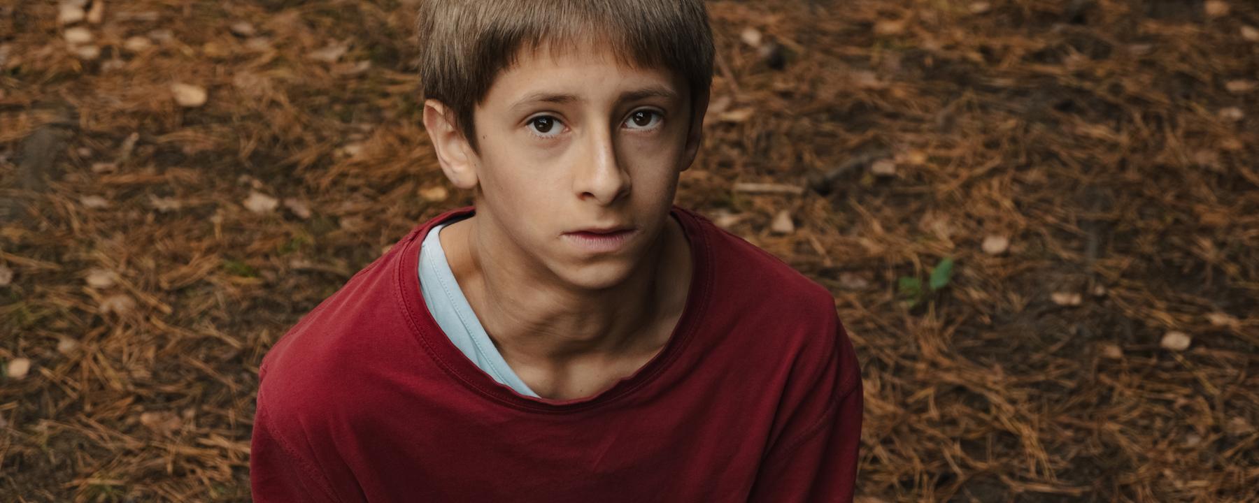  Man schaut von oben auf einen Jungen, ungefähr 12 Jahre, in einem rotem Pullover. Er schaut steht auf Waldboden und schaut ein wenig ängstlich und ernst nach oben. 
