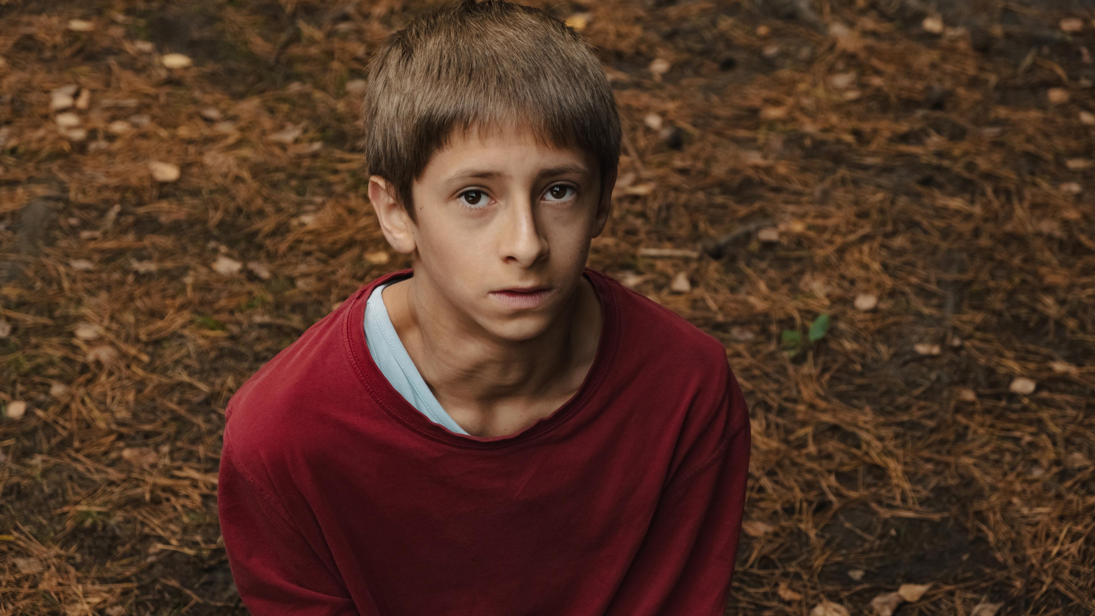  Man schaut von oben auf einen Jungen, ungefähr 12 Jahre, in einem rotem Pullover. Er schaut steht auf Waldboden und schaut ein wenig ängstlich und ernst nach oben. 