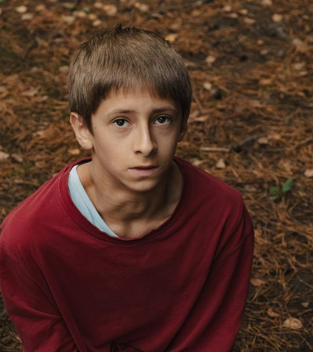  Man schaut von oben auf einen Jungen, ungefähr 12 Jahre, in einem rotem Pullover. Er schaut steht auf Waldboden und schaut ein wenig ängstlich und ernst nach oben. 